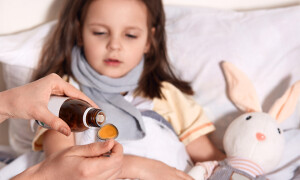Horizontal photo of unknown hand pouring liquid into spoon from little bottle with syrup, person caring about child lying in bed with flu and sore throat, being upset. Children and health concept.
